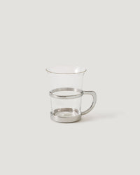 A clear glass mug with a polished silver metal band around the middle and a matching silver base, set against a solid light gray background.